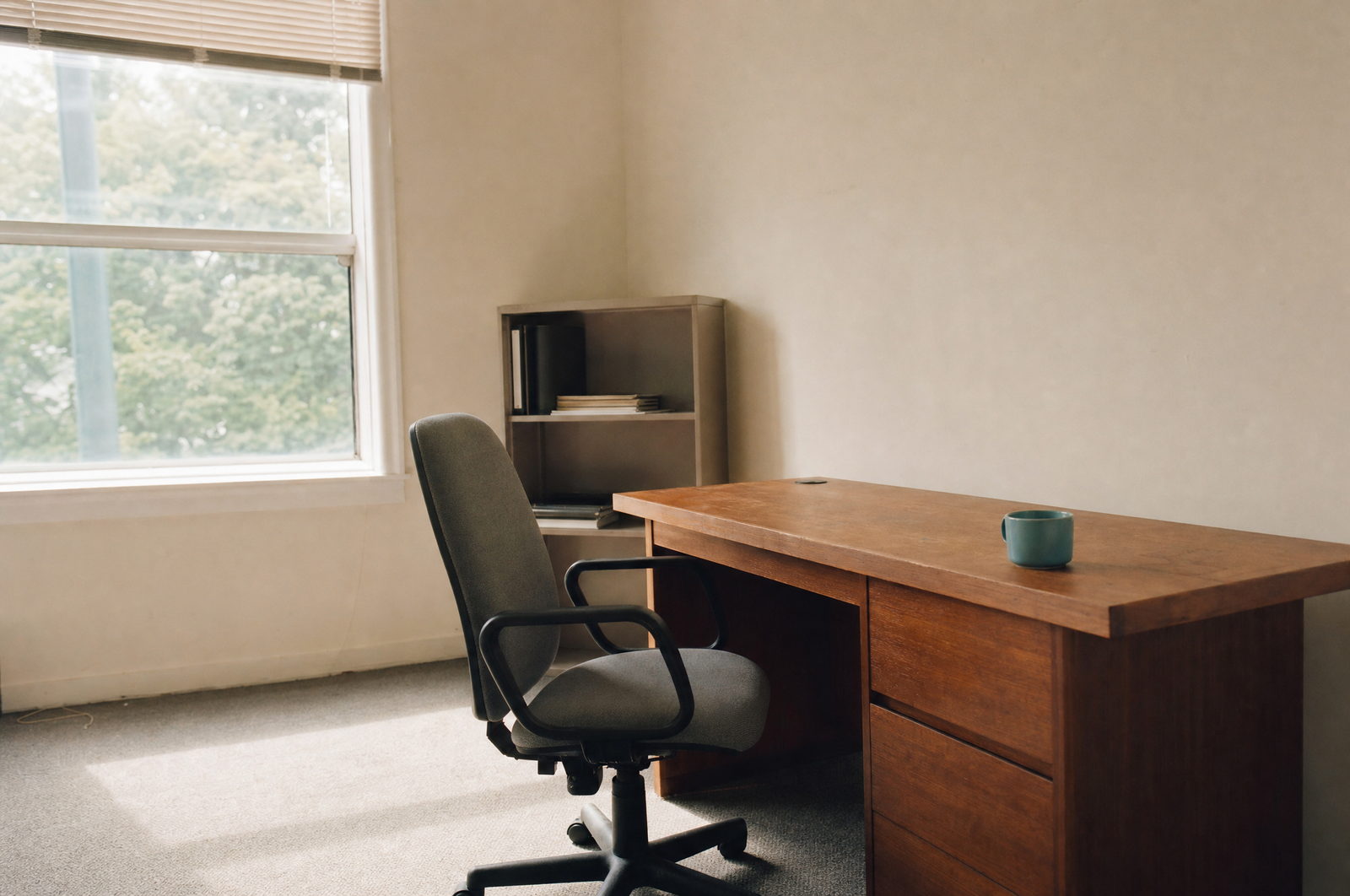 A quiet corner of a small office: a plain wooden desk, an empty task chair and a window with daylight falling across the floor.